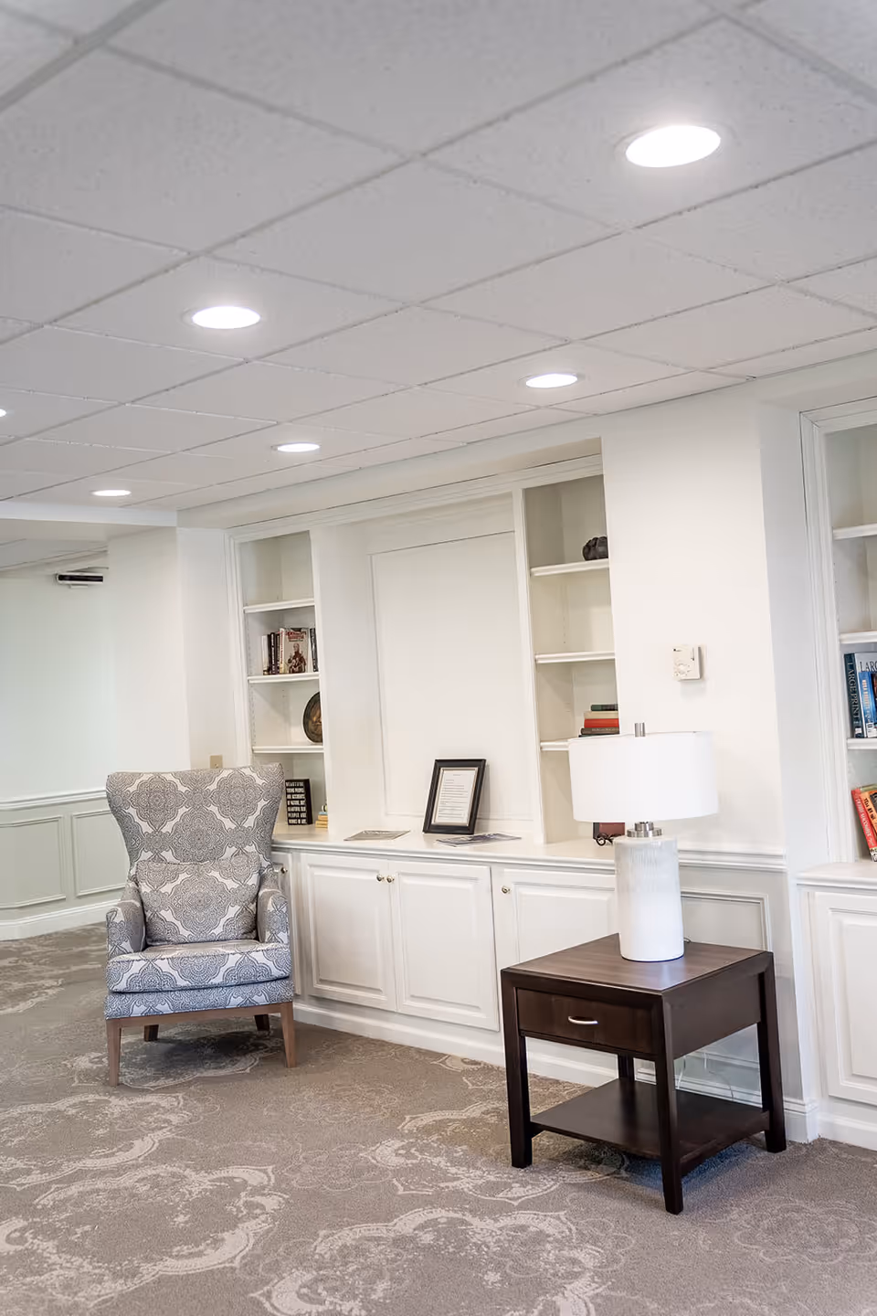 A cozy seating area in a senior living facility featuring a patterned armchair, a dark wooden side table with a white lamp, built-in white shelves with books and decorative items, and a carpeted floor with a subtle floral pattern.
