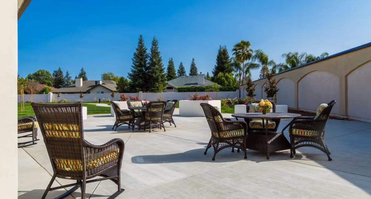 Outdoor patio area with several round tables and wicker chairs with cushions, surrounded by a concrete floor, greenery, and a white wall with trees and houses in the background under a clear blue sky.