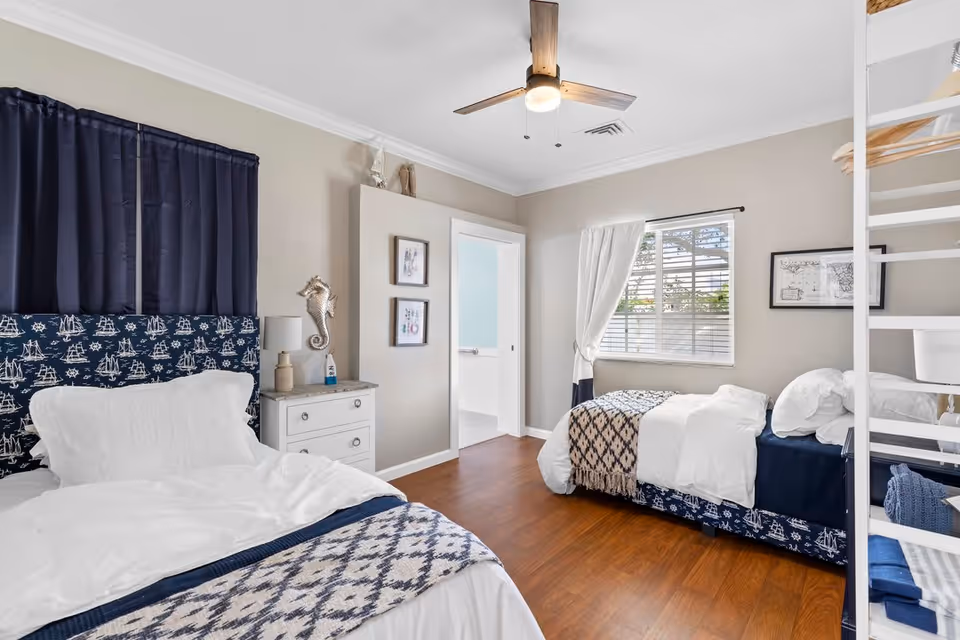 A bright bedroom with two beds featuring nautical-themed bedding and headboards. The room has wooden flooring, a ceiling fan with light, a window with white curtains, and a small white dresser with a lamp and decorative items. There is a door leading to a bathroom and a white ladder on the right side of the image.