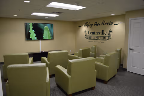 A small movie theater room with six green armchairs arranged in two rows facing a wall-mounted flat screen TV. On the wall above the chairs, there is a decorative sign that reads 'Enjoy the Movie Centreville Theater Please turn off your cell phone.' The room has a beige wall and a white door on the right side.