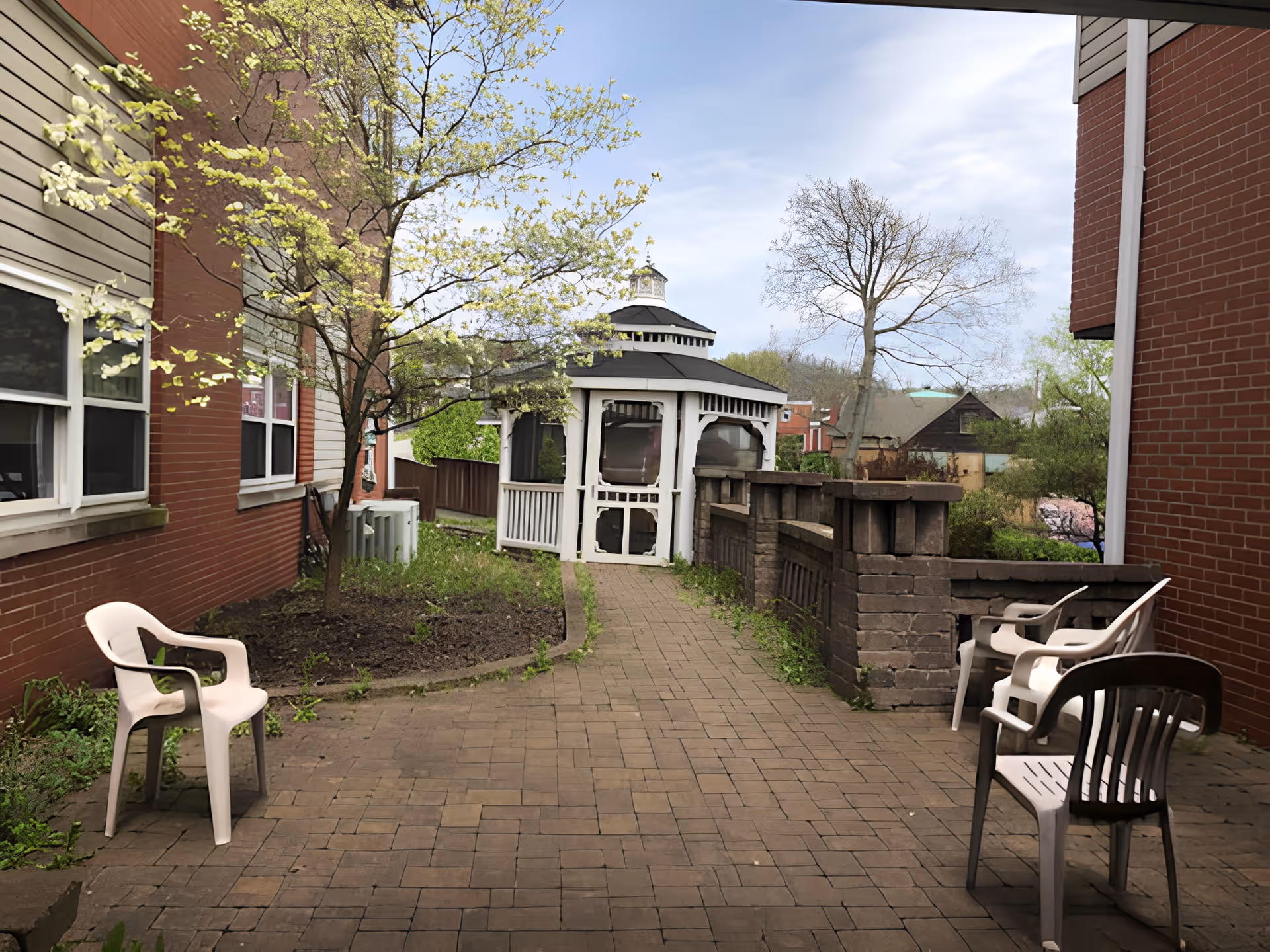 Outdoor patio area between two brick buildings with a paved walkway leading to a white gazebo. There are several plastic chairs placed around the patio, some white and one dark-colored. Trees with budding leaves and some greenery surround the area under a partly cloudy sky.