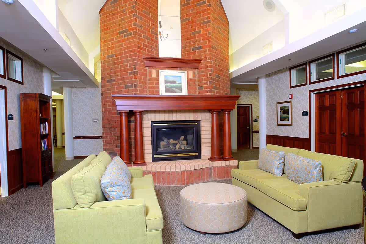 A cozy senior living common area with two light green sofas facing each other around a round beige ottoman. In the background, there is a large brick fireplace with wooden columns and a framed picture above it. The room has carpeted floors, wooden doors, and soft lighting.