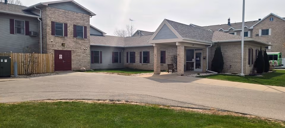 Exterior view of a single-story brick and siding building with a covered entrance, a driveway, and a small grassy area in front. The building has multiple windows with dark shutters and a bench near the entrance.