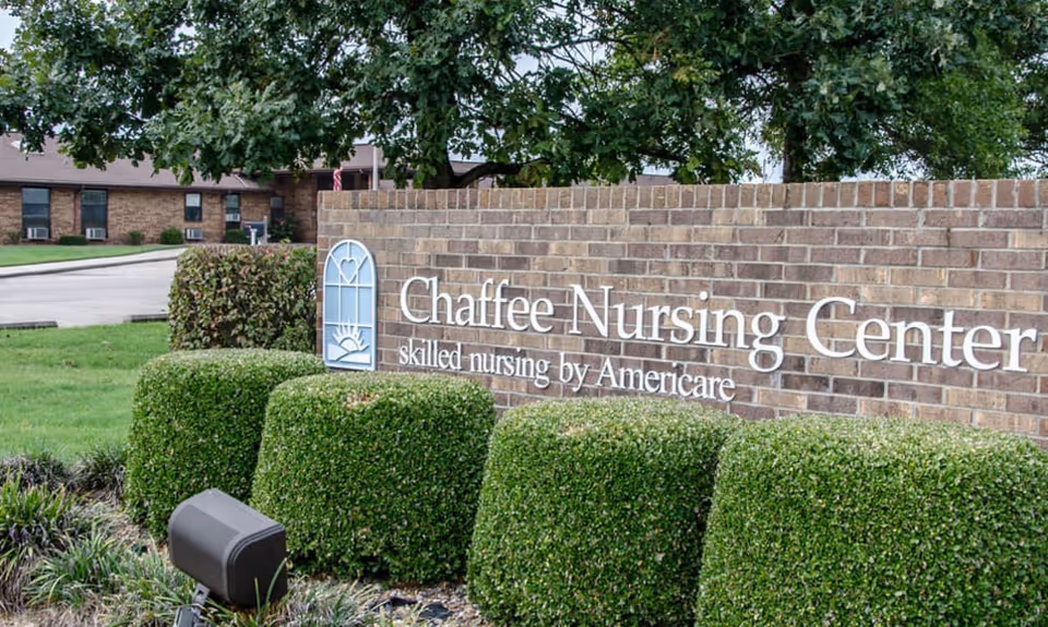 A brick sign for Chaffee Nursing Center with the text 'skilled nursing by Americare' surrounded by neatly trimmed green bushes and trees in the background.