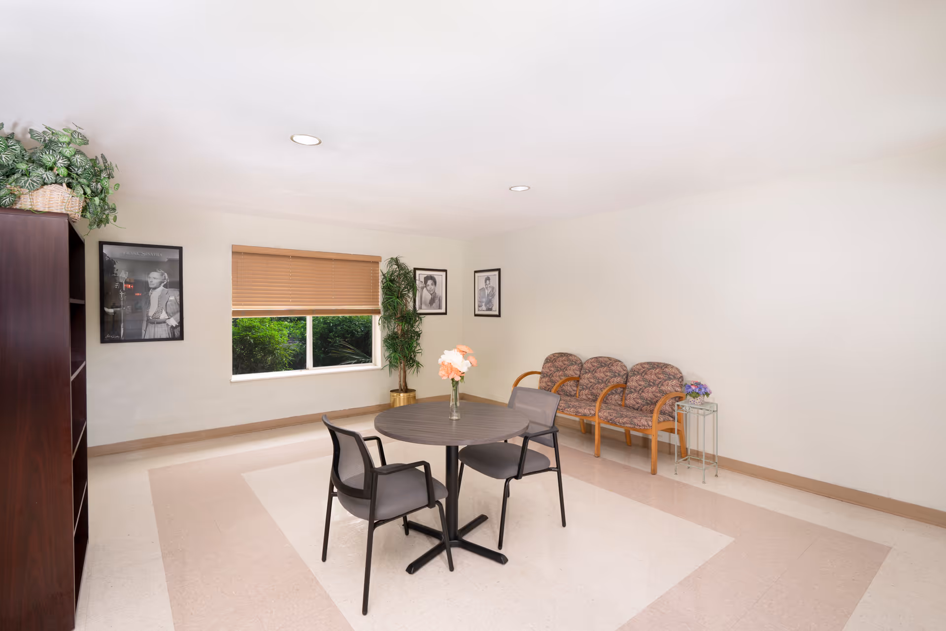 A small sitting area in a senior living facility with a round table and two chairs in the center. There are three cushioned chairs with wooden armrests against the wall on the right. A small side table with a flower vase is next to the chairs. A large window with closed blinds is on the back wall, flanked by two framed black and white photographs. A tall potted plant is placed near the window. A dark wooden bookshelf with a plant on top is on the left side of the room. The floor has a light-colored tile with a beige border, and the walls are painted white.