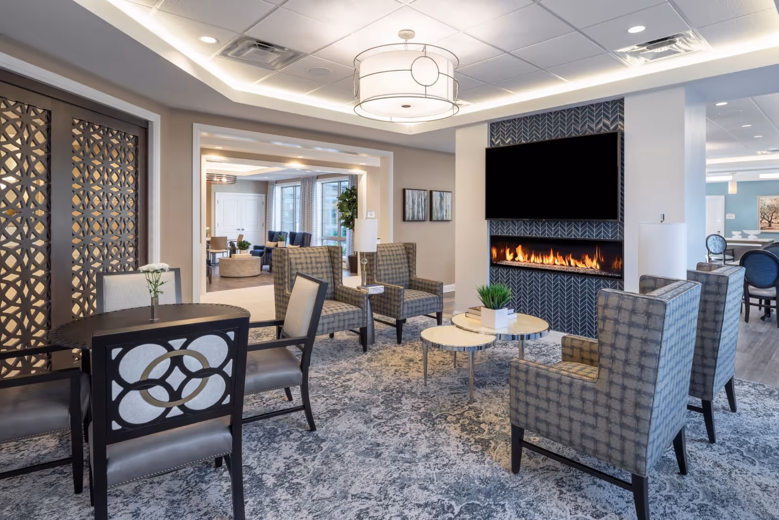 A modern and cozy living room area in a senior living facility featuring a patterned carpet, several upholstered armchairs arranged around a set of nested round tables with a small plant and books, a wall-mounted electric fireplace beneath a large flat-screen TV, and a round table with chairs in the foreground. The room is well-lit with ceiling lights and has decorative wall art and large windows in the background.