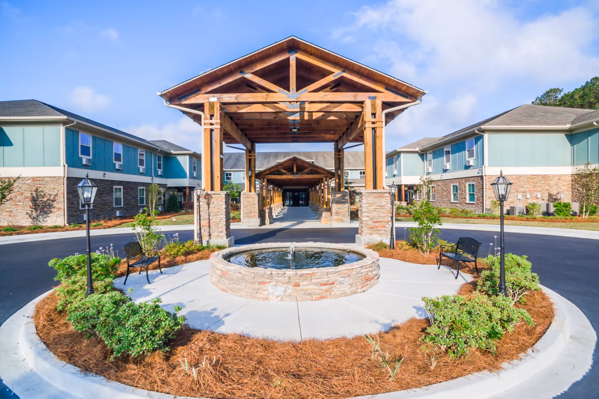 Exterior view of Oaks at Pooler senior living facility featuring a circular driveway with a stone fountain in the center, surrounded by landscaped bushes and benches. The building has two stories with a combination of stone and teal siding, and a wooden covered entrance with exposed beams.