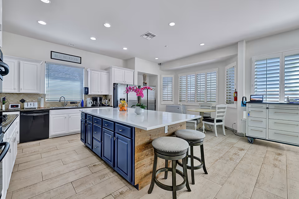 Bright and spacious kitchen with white cabinets and a large island featuring blue cabinetry and a white countertop. Two cushioned stools are placed at the island. The kitchen has stainless steel appliances, a window with blinds, and a sign above the window that reads 'Bless This Kitchen'. In the background, there is a small dining area with a round table and chairs, and large windows with white shutters allowing natural light to fill the room.
