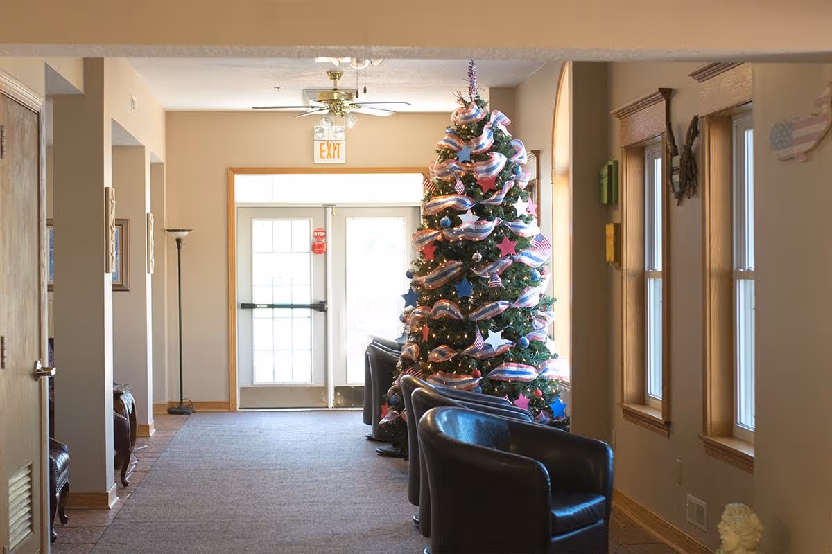 Interior lobby hallway with a decorated Christmas tree and black chairs along a carpeted walkway leading to an exit door.