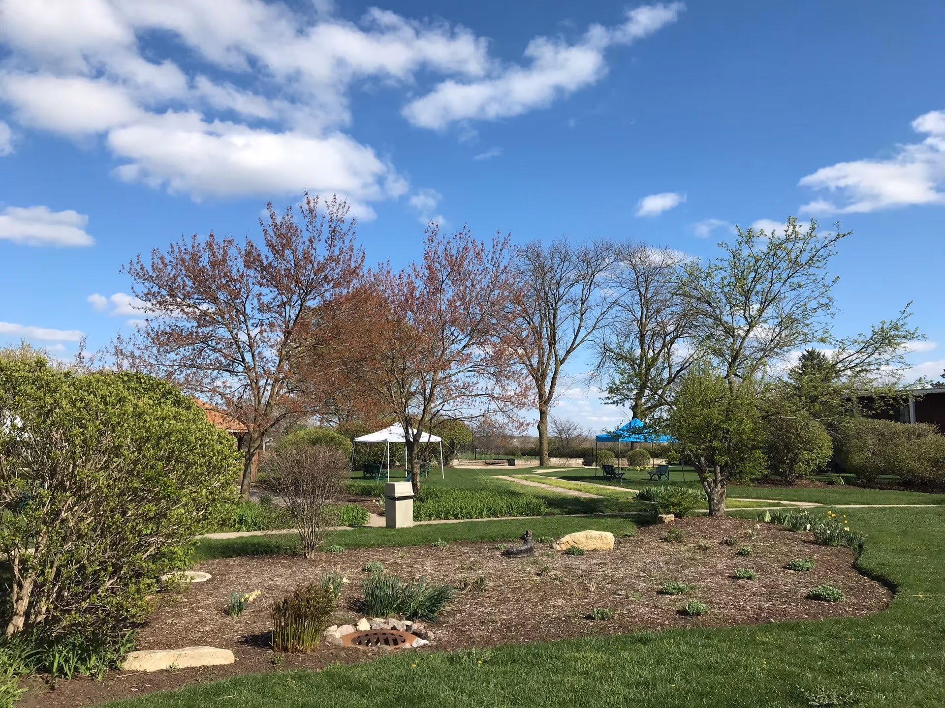 A landscaped outdoor garden area with green grass, bushes, and trees under a blue sky with scattered clouds. There are two canopy tents, one white and one blue, with benches underneath them along a paved walking path.