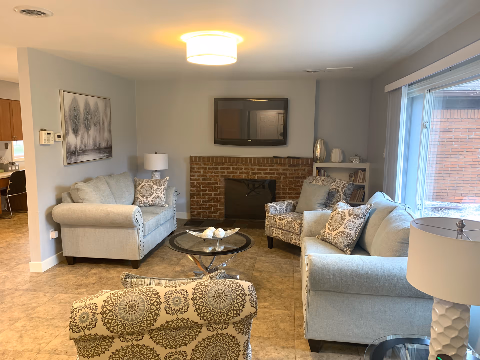 A cozy living room with two light gray sofas, two patterned armchairs, a round glass coffee table, a brick fireplace with a mounted flat-screen TV above it, a side table with a lamp, a bookshelf, and a large window letting in natural light.