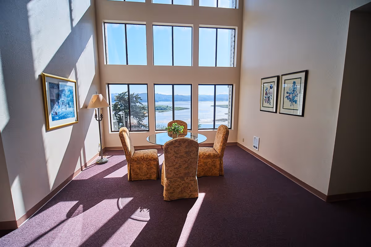 A bright room with large windows offering a scenic view of a body of water and distant hills. The room contains a round glass table surrounded by four upholstered chairs with patterned fabric. There is a floor lamp and framed artwork on the walls, and sunlight casts shadows on the purple carpeted floor.