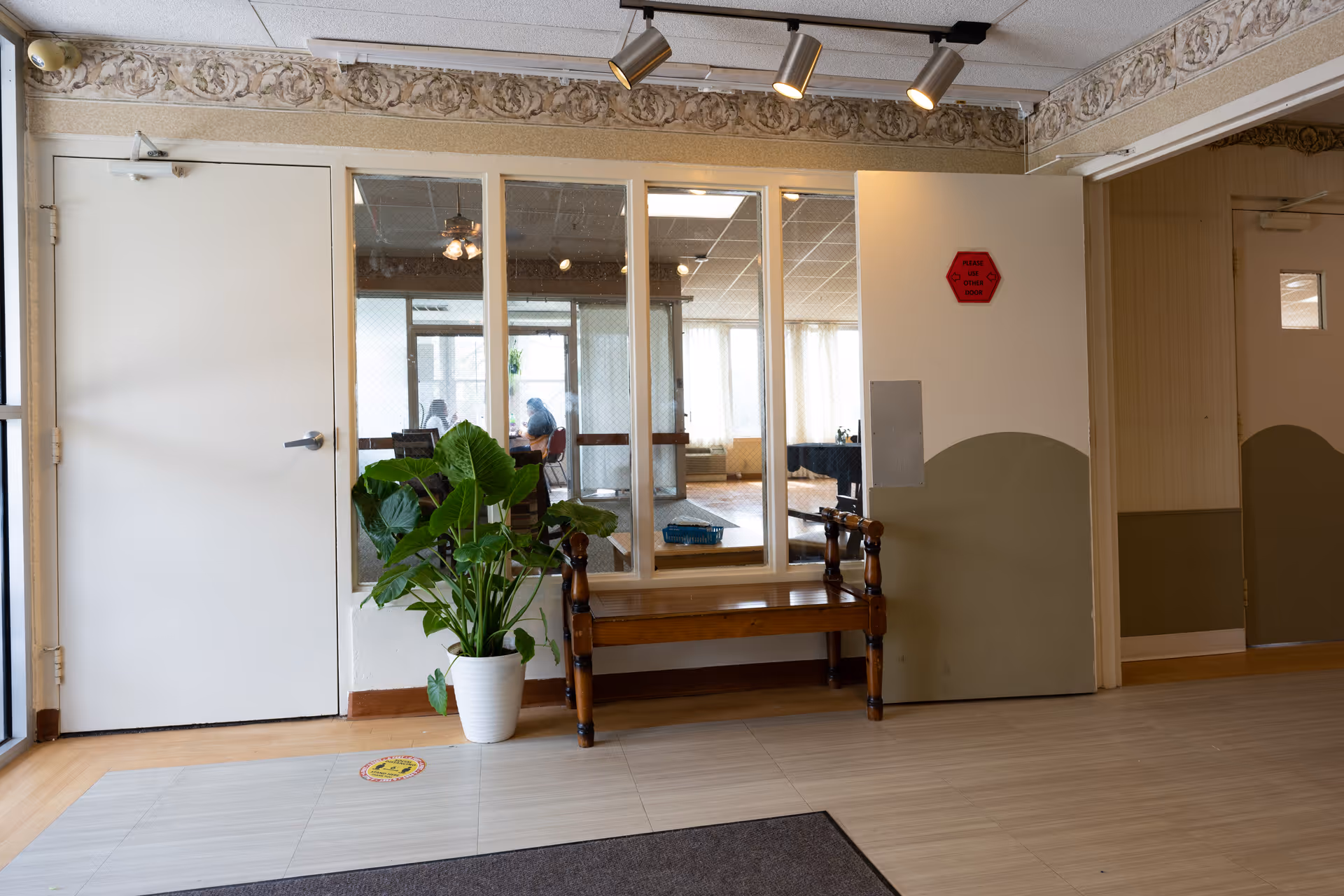 Interior view of a hallway in a senior living facility with a wooden bench, a potted green plant, and a set of windows looking into a room where people are seated. The walls are painted white and olive green with a decorative border near the ceiling. There are two closed doors, one white and one beige, and ceiling spotlights illuminating the area.