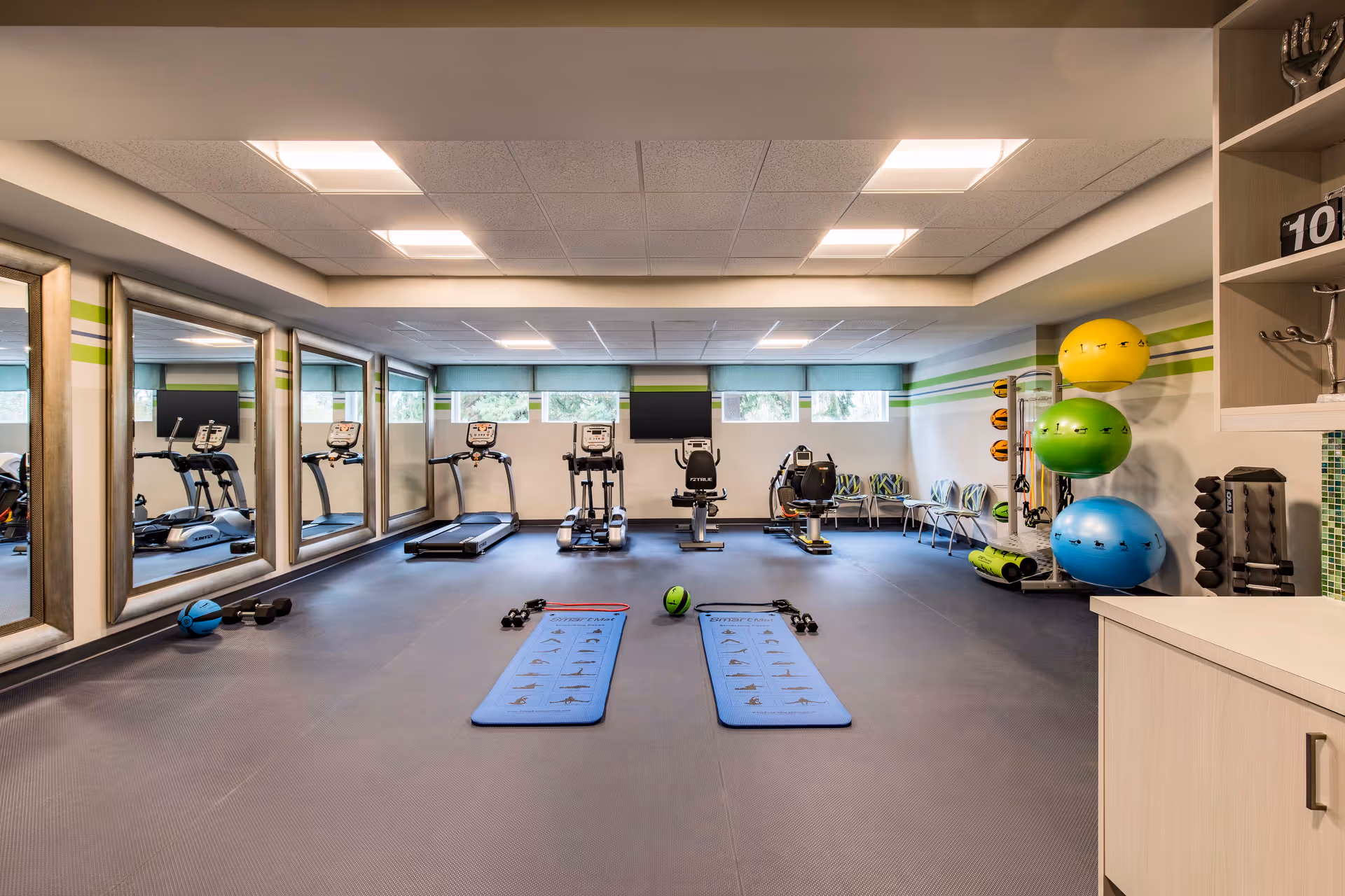 A well-lit fitness room with exercise equipment including treadmills, stationary bikes, dumbbells, exercise mats, and large colorful exercise balls. The room has large mirrors on one wall, windows on the opposite wall, and chairs lined up along the back wall.