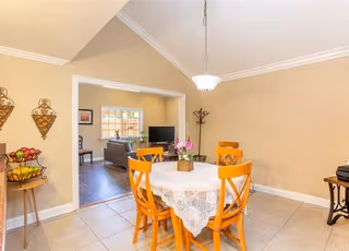 A dining area with a round table covered by a white lace tablecloth and four wooden chairs. A small flower arrangement is placed in the center of the table. The room has beige walls and a tiled floor. A hanging light fixture is above the table. In the background, there is an open doorway leading to a living room with a sofa and a window.