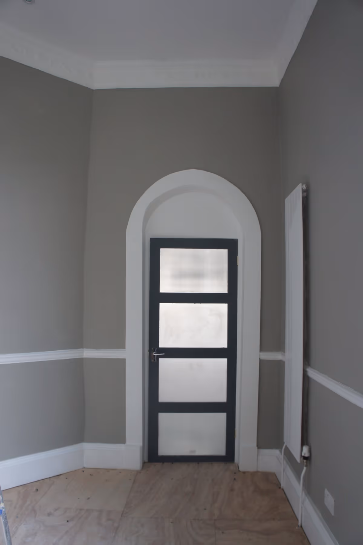 A small interior room with gray walls and white trim, featuring a black-framed door with frosted glass panels set within an arched white doorway. The floor appears unfinished with plywood sheets, and a white radiator is mounted on the right wall.