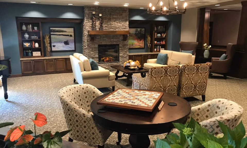 A cozy living room area with a stone fireplace in the center, flanked by built-in wooden shelves and cabinets. The room features comfortable seating including patterned armchairs, cream sofas, and a round table with a Scrabble board game on it. There are decorative plants and framed artwork on the walls, creating a warm and inviting atmosphere.