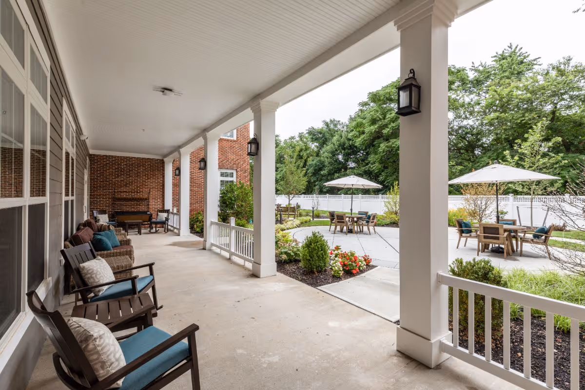 Covered outdoor patio area with several cushioned chairs and a bench along the wall, overlooking a landscaped garden with tables and chairs under umbrellas, surrounded by greenery and a white fence.
