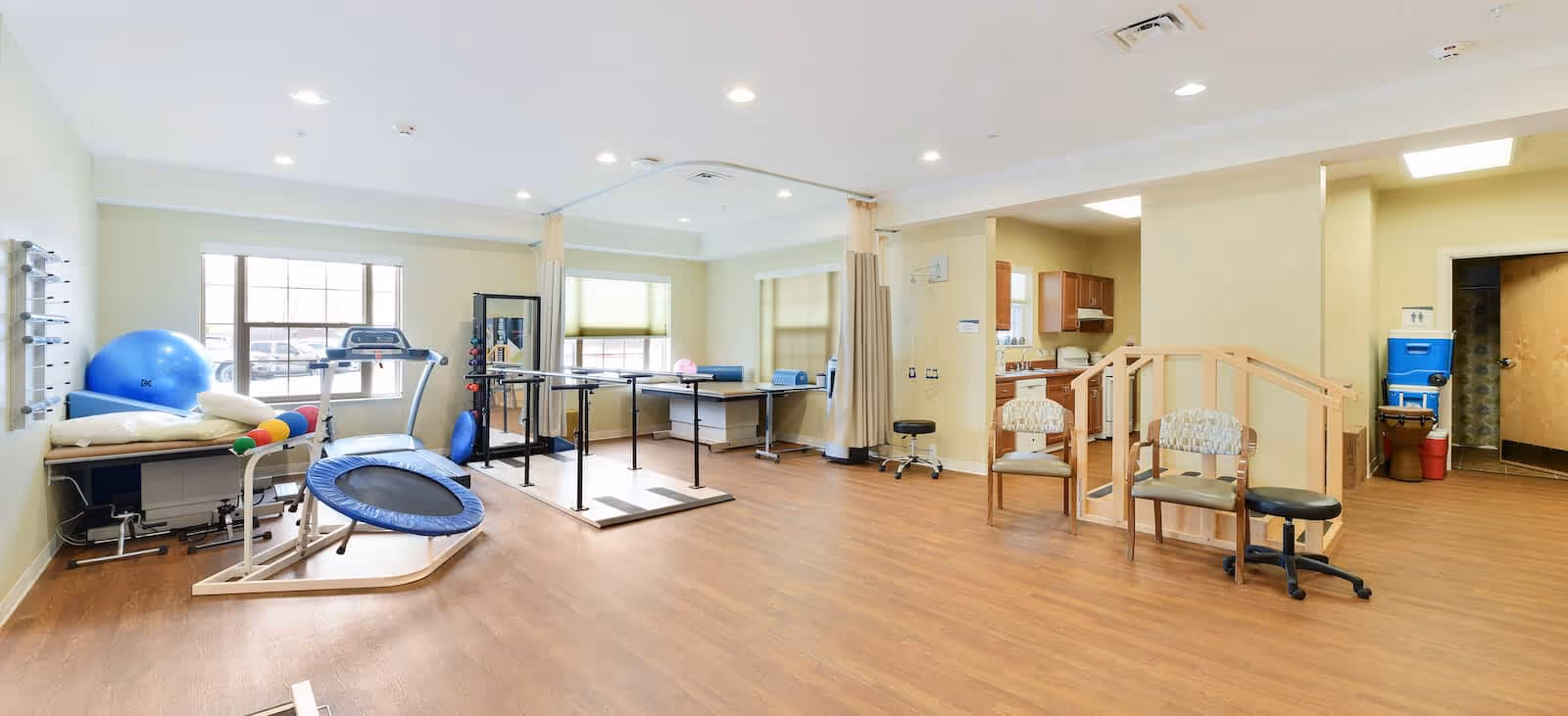 A spacious physical therapy room with wooden flooring and light yellow walls. The room contains various exercise equipment including a treadmill, a small trampoline, therapy balls, parallel bars for walking practice, and padded tables. There are two chairs and a small kitchen area visible in the background. Large windows allow natural light to fill the room.