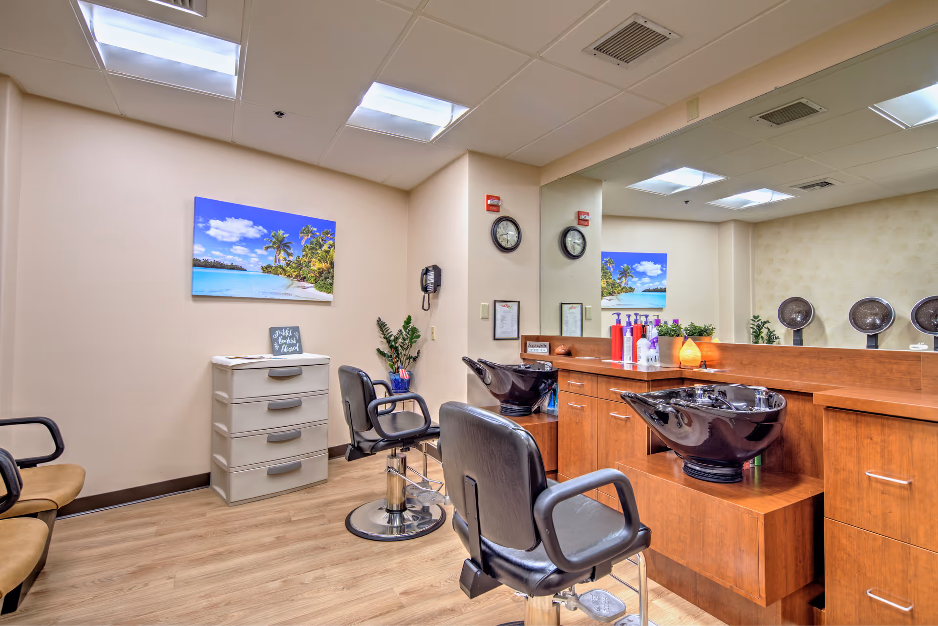 Interior of a hair salon area with two black salon chairs in front of a wooden counter with two black wash basins. The counter has various hair care products and a small lamp. There is a large mirror on the wall behind the counter, reflecting the room. On the left side, there is a white drawer unit, a black chair, and a wall-mounted phone. A tropical beach landscape picture hangs on the wall above the drawer unit. The floor is wooden, and the ceiling has fluorescent lights.