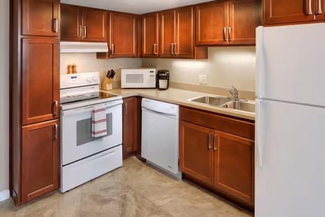 A clean kitchen with wooden cabinets, a white stove with a towel hanging on the handle, a microwave, a coffee maker, a dishwasher, a double sink, and a white refrigerator.