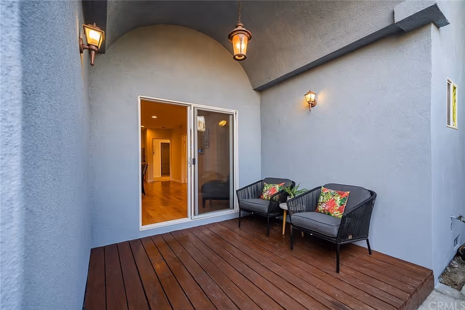 A small outdoor patio area with wooden flooring, two black wicker chairs with gray cushions and colorful floral pillows, a small side table with a plant, and three wall-mounted lantern-style lights. The patio is adjacent to a building with light gray stucco walls and a sliding glass door leading inside.