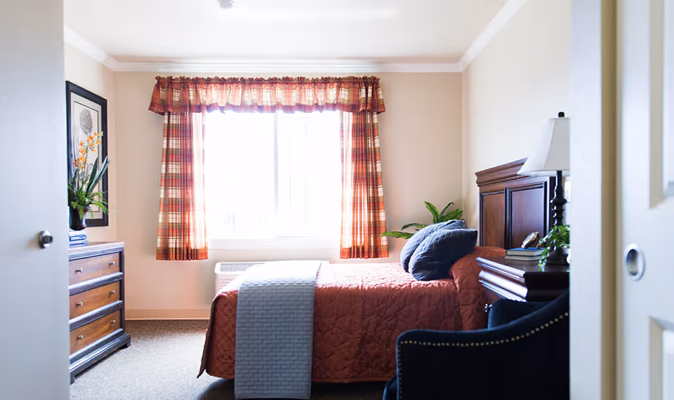 A cozy bedroom with a bed covered in a rust-colored quilt and a gray throw blanket. The bed has two dark pillows and is positioned against a wall with a wooden headboard. A wooden dresser with a plant and framed artwork is on the left side of the room. A window with plaid curtains lets in natural light, and a dark upholstered chair is partially visible in the foreground.