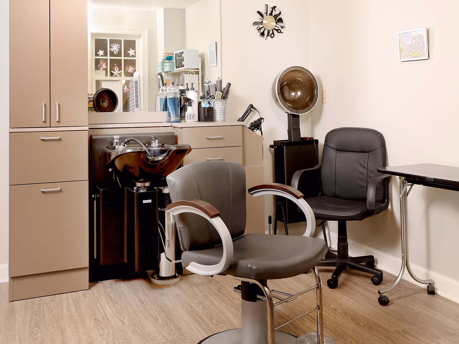 Interior of a hair salon area with a black hair washing sink, a gray salon chair with wooden armrests, a black rolling chair, a hair dryer hood, and various hair care products on a countertop with cabinets. The walls are light-colored with a clock and framed artwork.