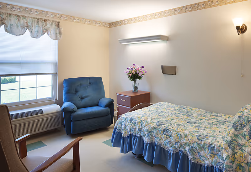 A cozy bedroom in a senior living facility with a single bed covered in a floral bedspread, a wooden nightstand with a vase of flowers, a blue upholstered armchair, and a window with blinds and floral valance letting in natural light.