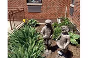 Two small statues of children, one boy and one girl, standing in a garden bed with green plants and flowers, next to a brick building with windows.