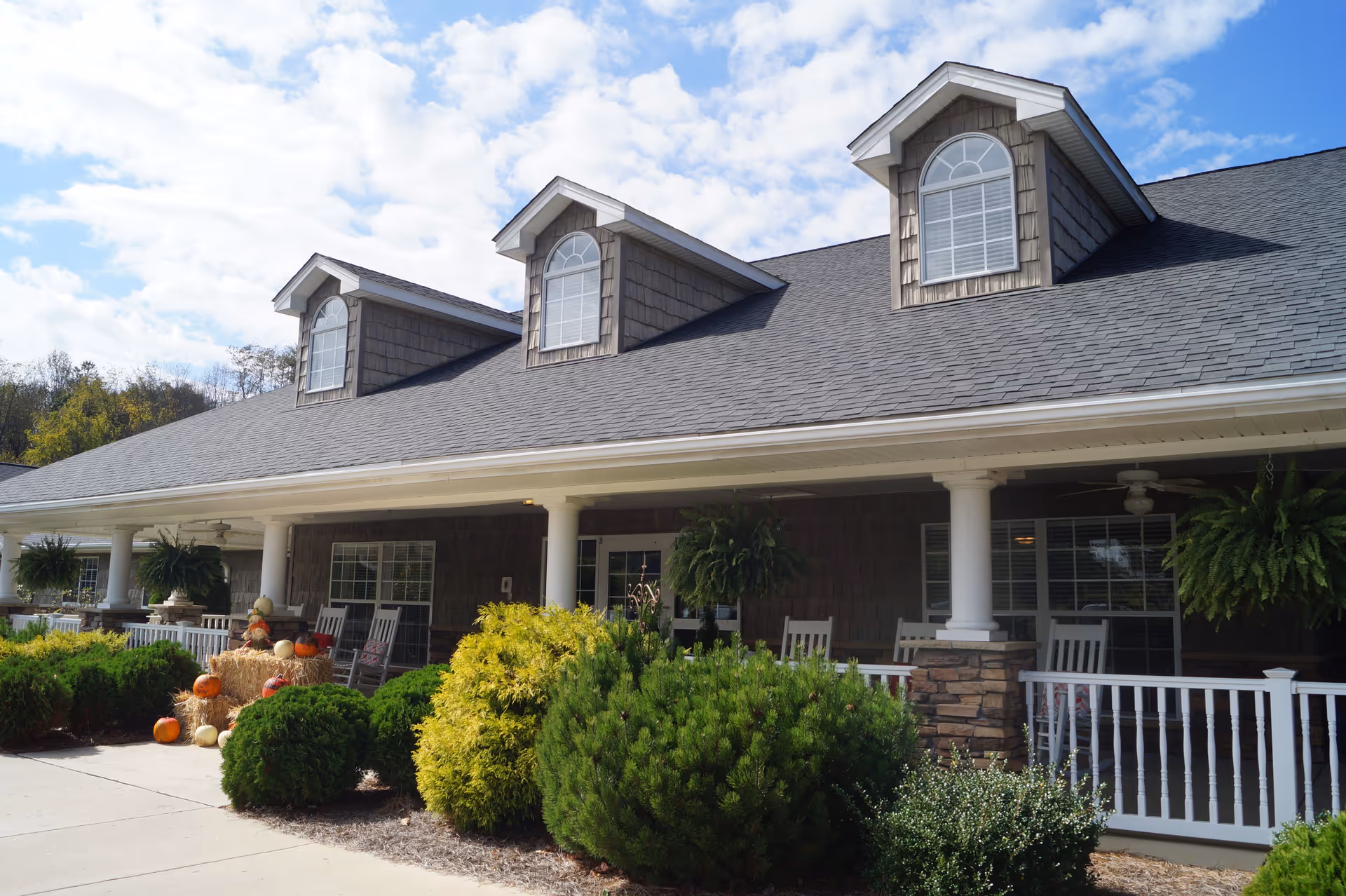 Exterior view of a senior living facility with a large covered porch featuring white rocking chairs, hanging ferns, and autumn decorations including pumpkins and hay bales. The building has a gray shingled roof with three dormer windows and white columns supporting the porch roof. Green shrubs and bushes line the walkway in front of the porch under a partly cloudy sky.