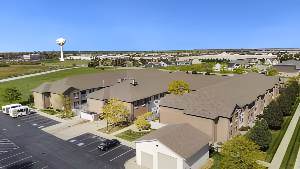 Aerial view of a large senior living facility building with a brown roof and beige walls, surrounded by green lawns and trees. There is a parking lot with a few vehicles and a water tower visible in the background under a clear blue sky.