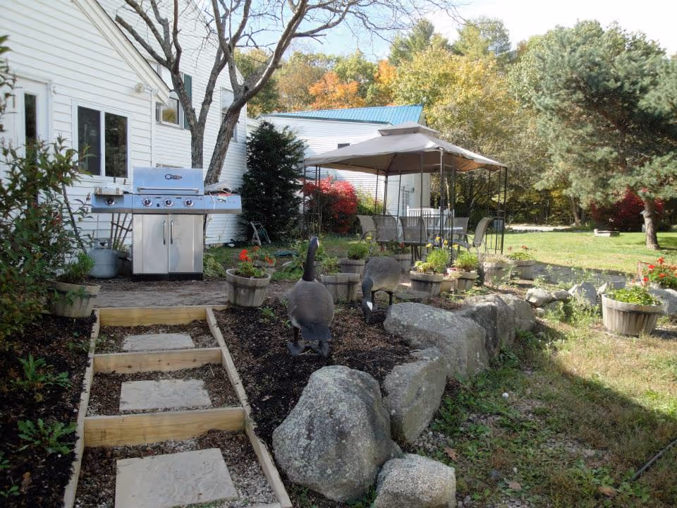 Outdoor garden area with a stone pathway leading to a patio covered by a canopy. The patio has a table and chairs. There are decorative goose statues, potted plants, large rocks, and a stainless steel barbecue grill next to a white building. Trees with autumn foliage are visible in the background.