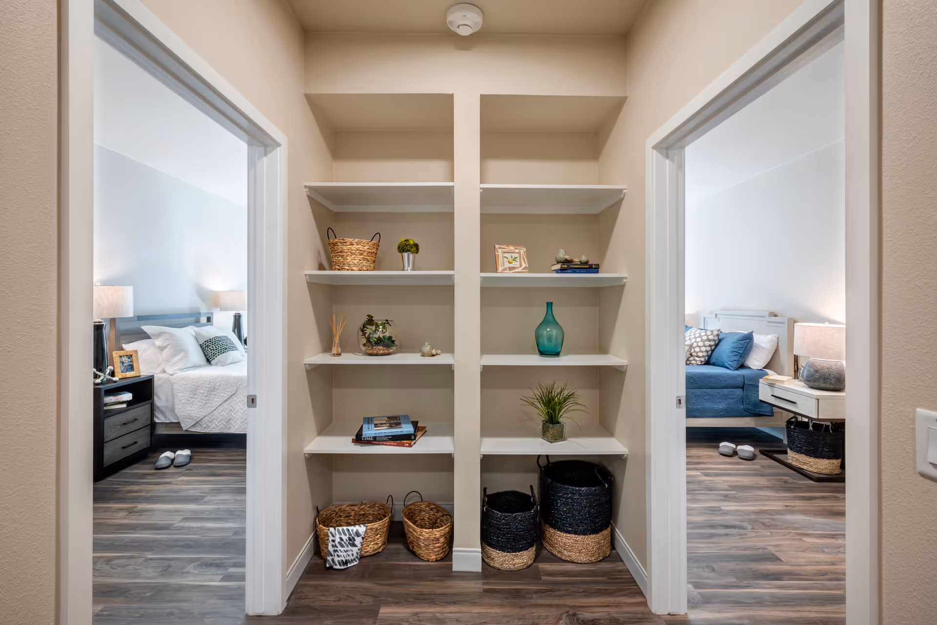 A hallway with built-in white shelves holding decorative items such as baskets, plants, books, and vases. On either side of the hallway are doorways leading to two bedrooms, each with a bed, nightstand, lamp, and slippers on the floor. The flooring is wood-style laminate and the walls are painted light beige.