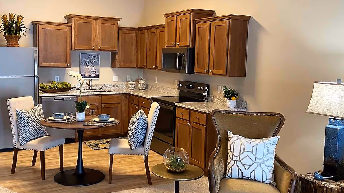 Open-concept kitchen with wooden cabinets, stainless appliances, a small round dining table set for two, and an upholstered armchair with a lamp.