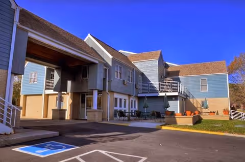 Exterior view of a two-story assisted living facility building with blue siding and brown roof shingles under a clear blue sky. There is a covered entrance supported by columns, outdoor seating with tables and umbrellas, and a handicapped parking space in the foreground.