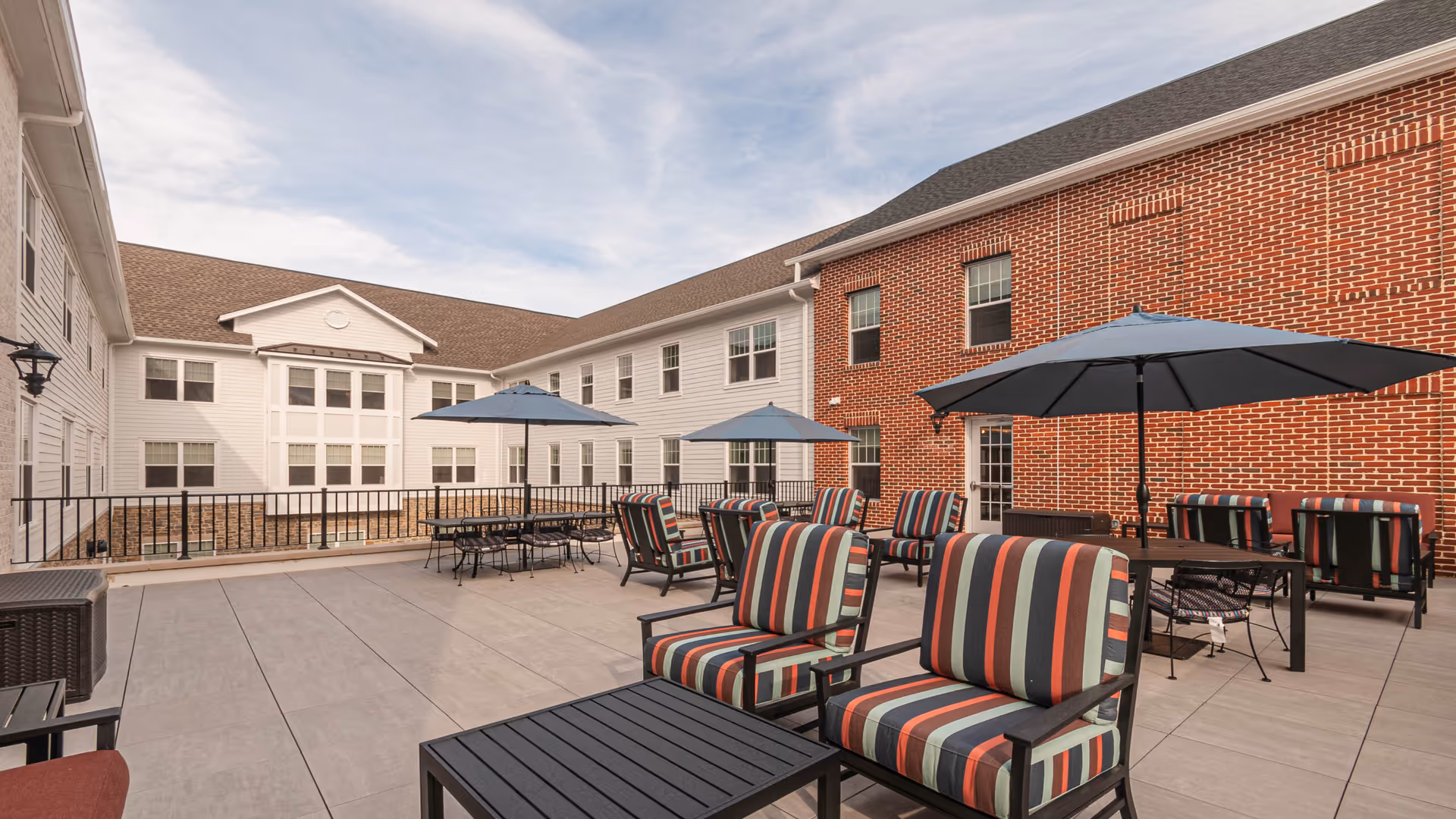 Outdoor patio area at Village Crossing at Worman's Mill featuring multiple seating arrangements with striped cushioned chairs and tables shaded by large blue umbrellas, surrounded by a two-story building with white and red brick exterior walls under a partly cloudy sky.