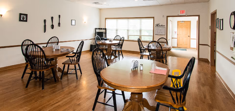A dining room with several round wooden tables and black wooden chairs arranged neatly on a wooden floor. The room has white walls with minimal decorations, a large window letting in natural light, and an exit door in the background. A sign on the wall near the door reads 'Kitchen'.