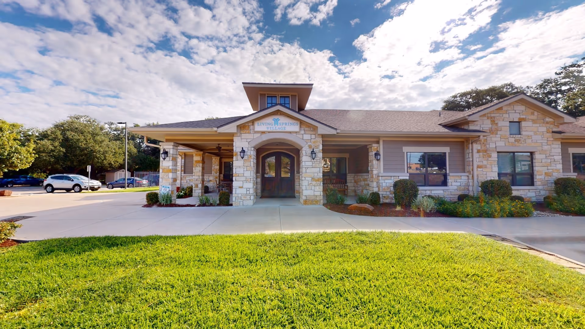 Front exterior view of Mercy House Living Springs building with stone and siding facade, a covered entrance with double doors, landscaped bushes, green grass lawn, parked cars, and a partly cloudy sky.