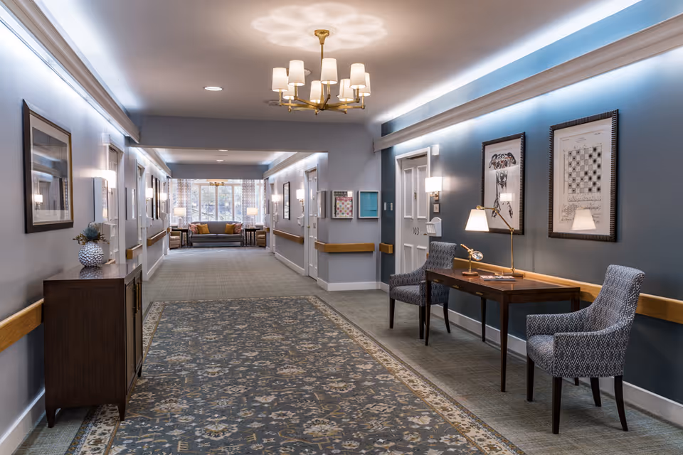 Well-lit furnished hallway with a patterned runner carpet, seating area at the far end, framed artwork, and a small desk with chairs along the wall.