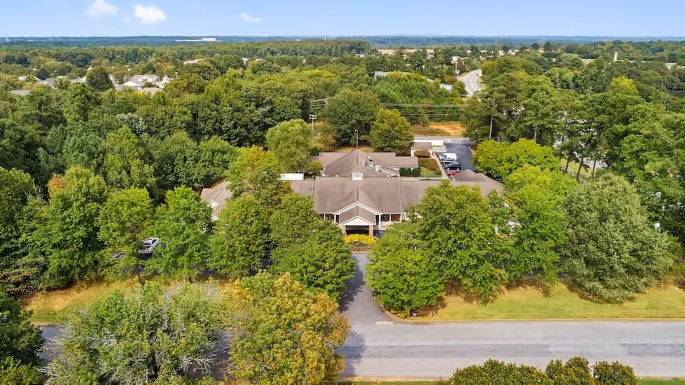 Aerial view of Spring Arbor Assisted Living facility surrounded by dense green trees and vegetation, with a road in the foreground and a parking area visible behind the building.