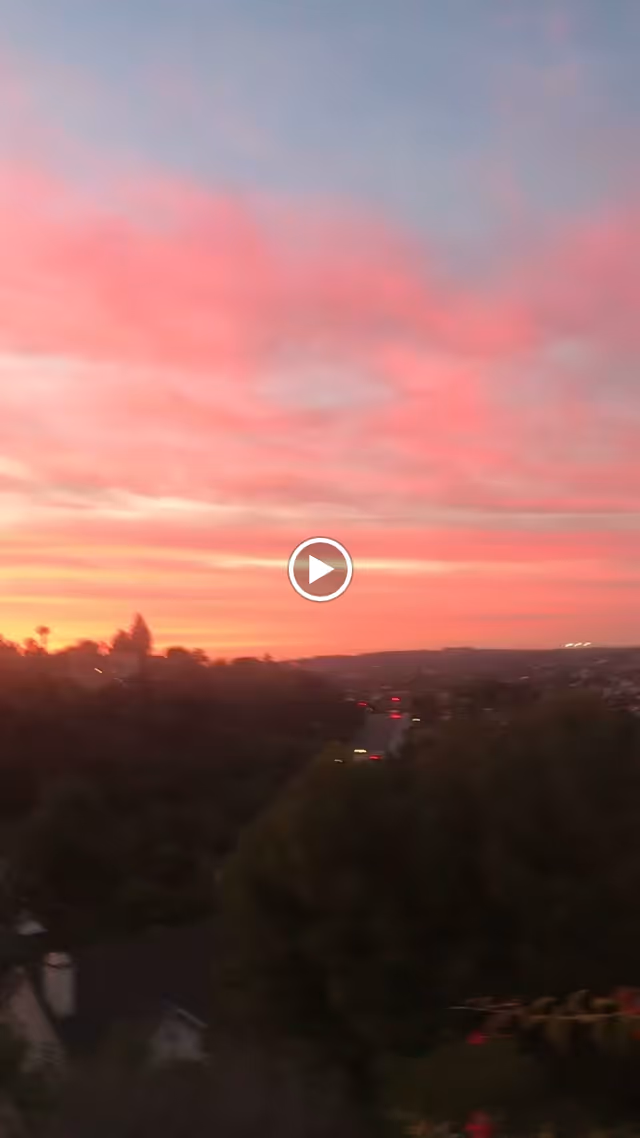 A scenic view of a sunset with a sky filled with pink and orange clouds over a landscape of trees and distant hills, with a road and some houses visible below.