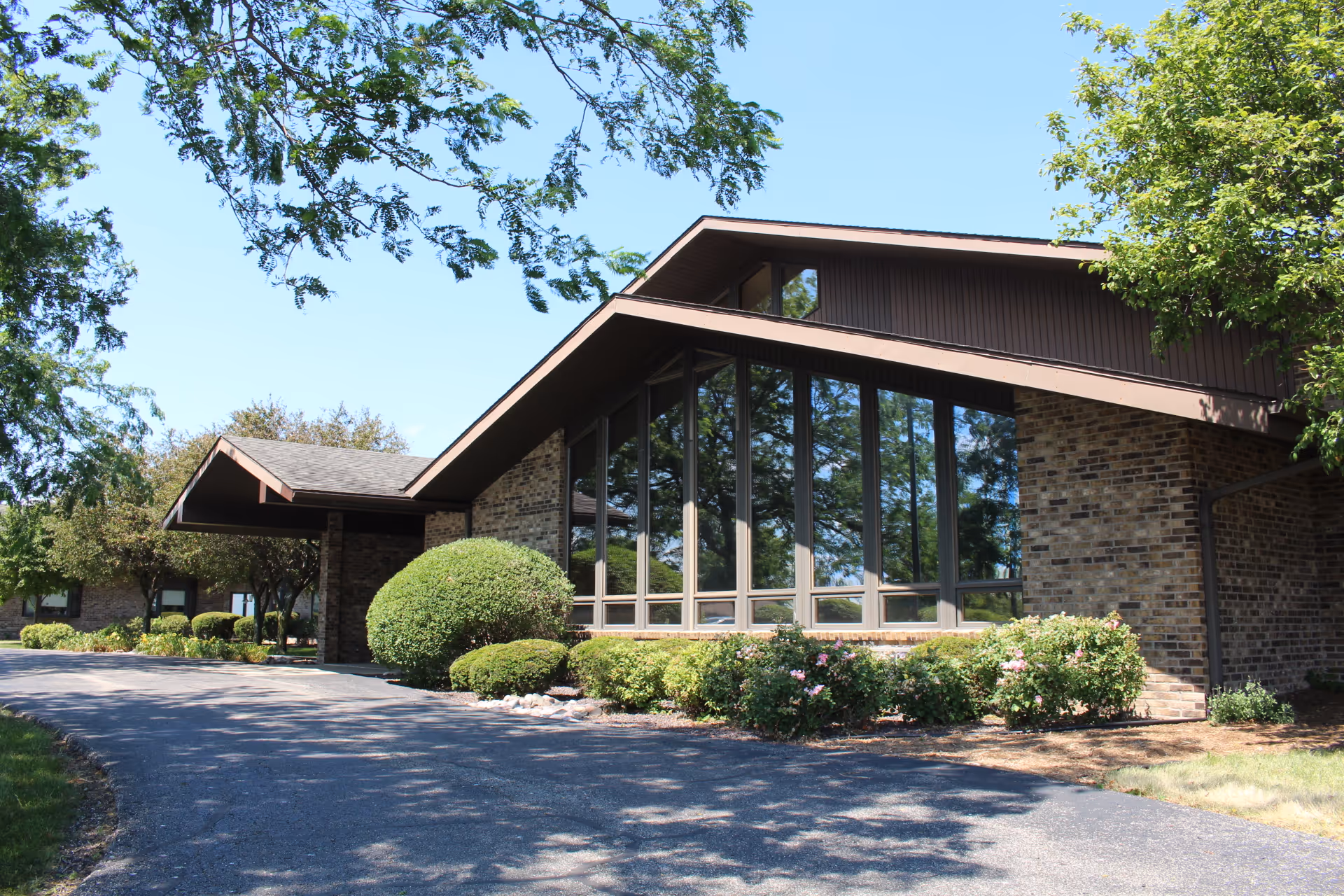 Exterior view of a single-story brick building with large triangular windows, surrounded by bushes and trees under a clear blue sky.
