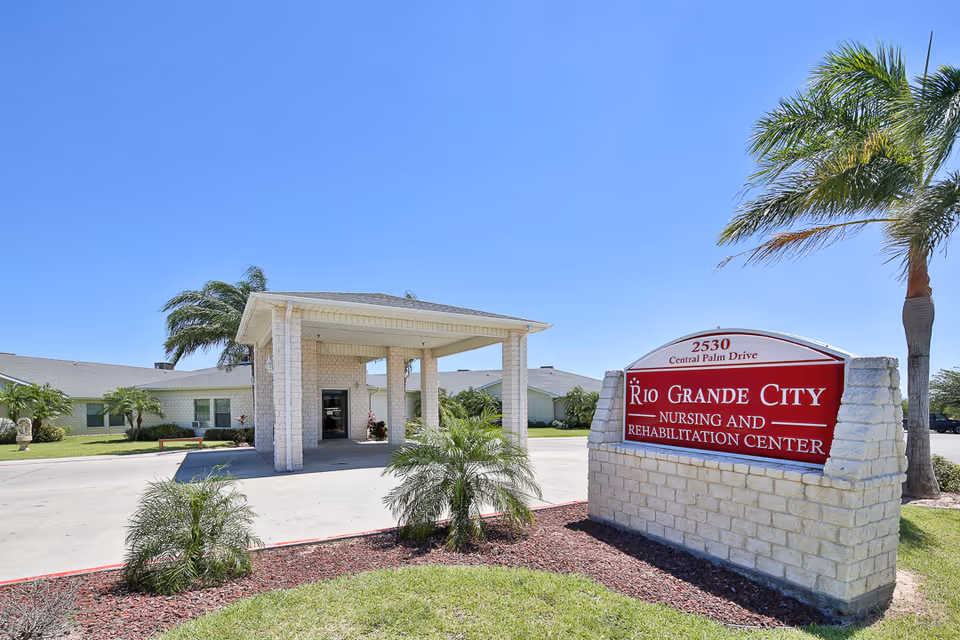 Exterior view of Rio Grande City Nursing and Rehabilitation Center building with a covered entrance, palm trees, and a red and white sign displaying the facility's name and address under a clear blue sky.