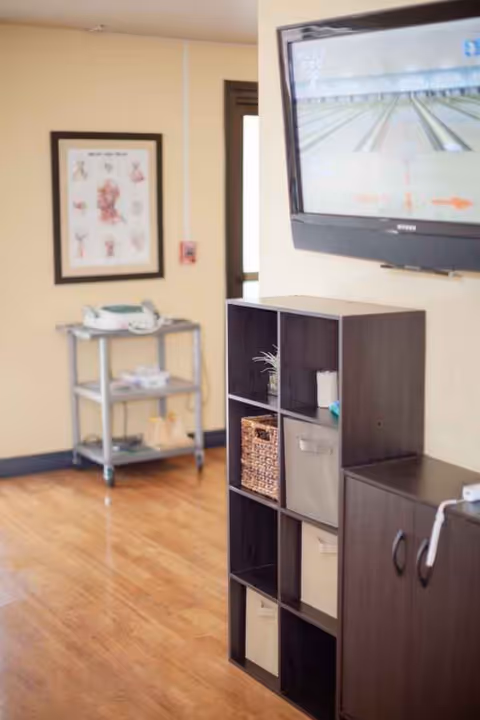 Interior of Lincoln Meadows Care Center shows shelving unit, wall-mounted TV, and medical cart.