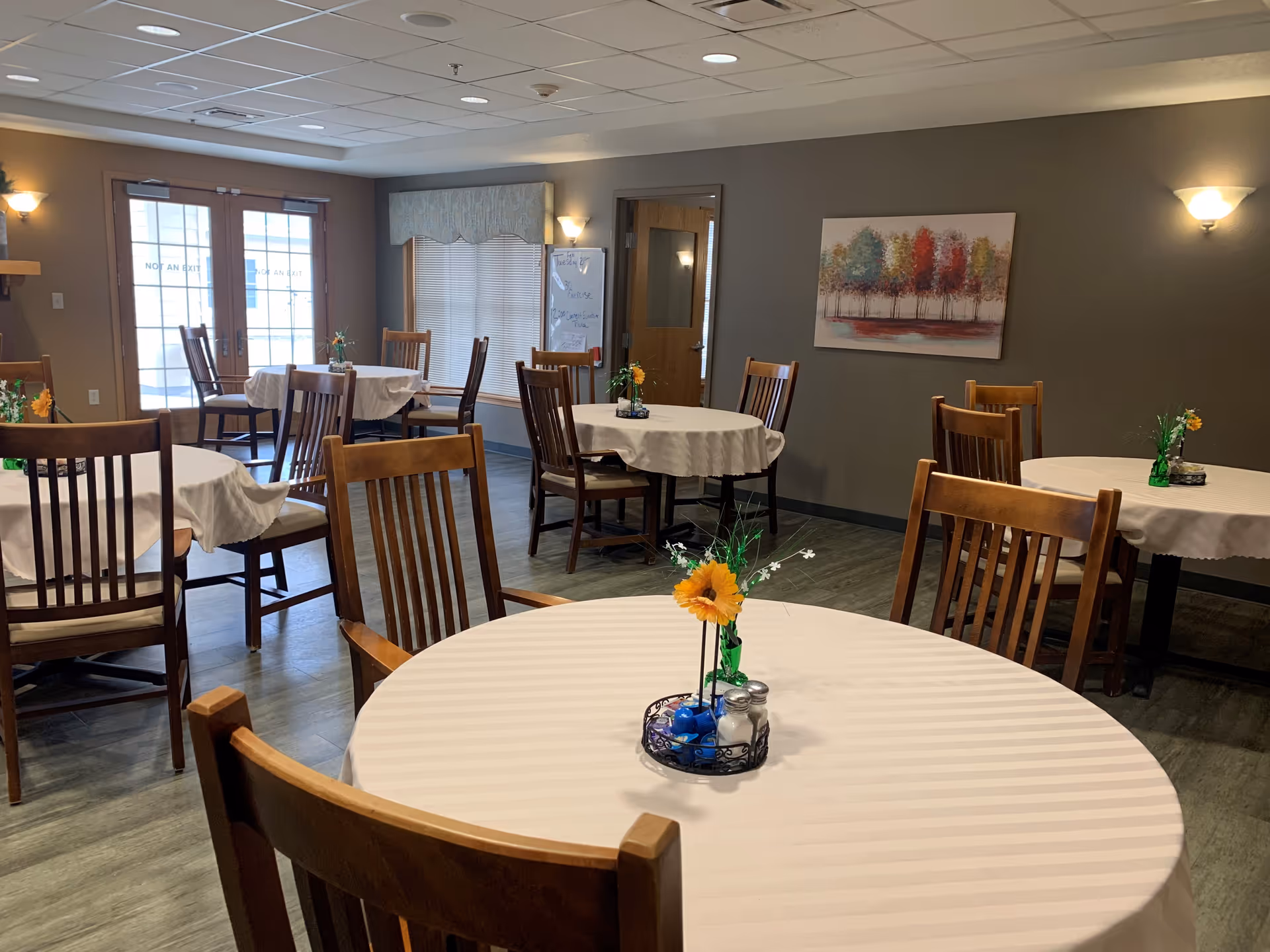 A dining room with several round tables covered with white tablecloths, each table decorated with a small flower arrangement. Wooden chairs surround the tables. The room has wood flooring, beige walls, and a painting of colorful trees on one wall. There are windows and a door with glass panels letting in natural light.