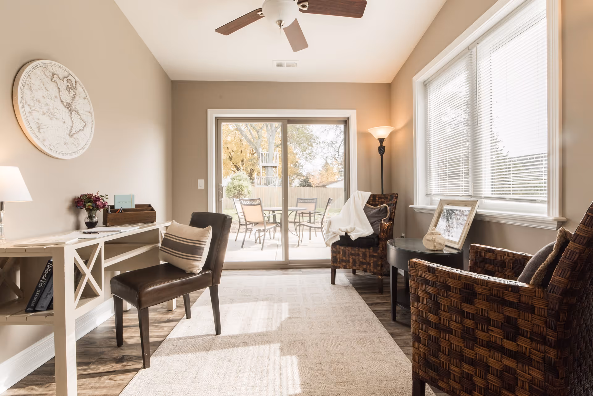Sunlit living room with a desk, leather and wicker chairs, and a sliding glass door leading to an outdoor patio.