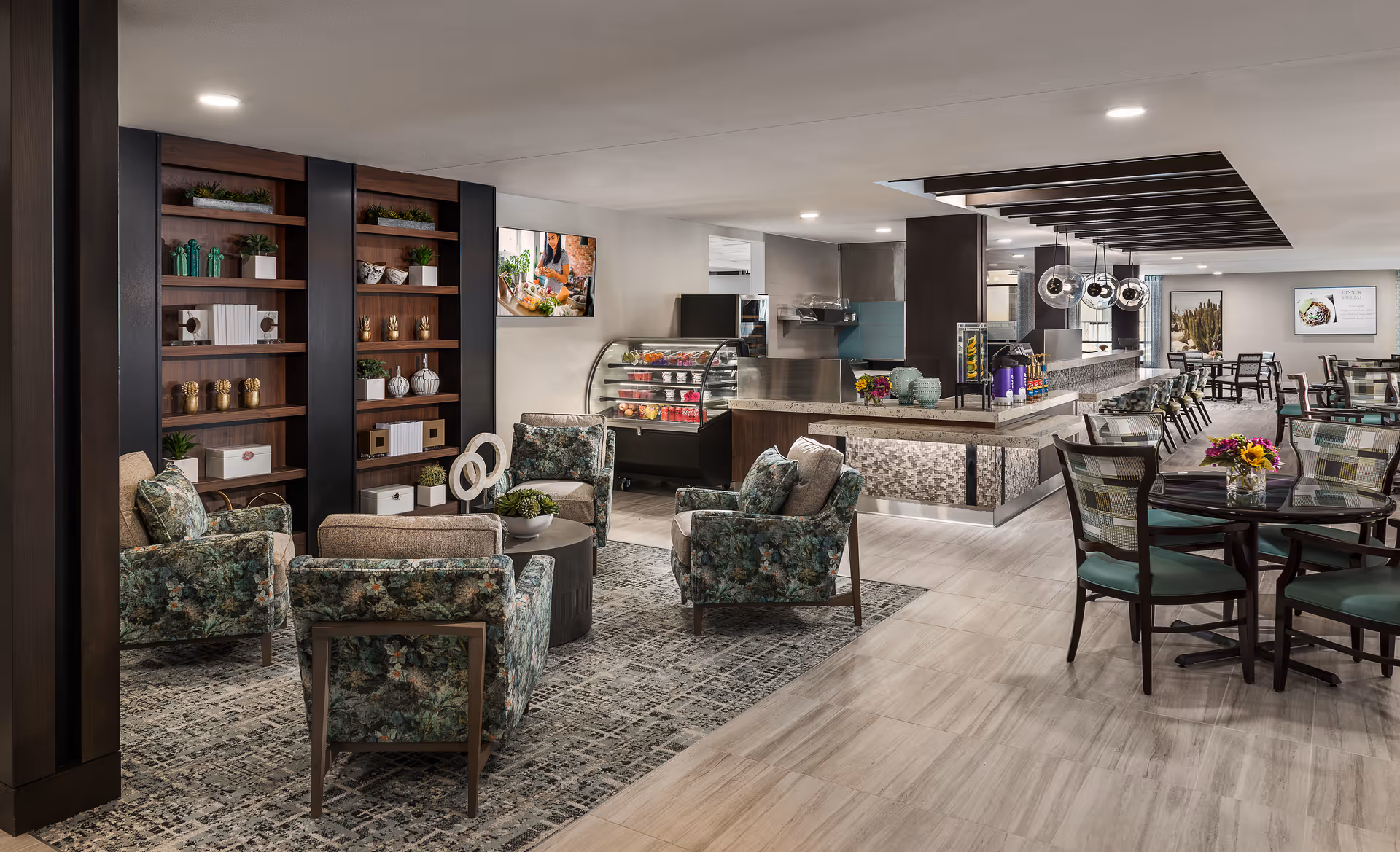 Open communal dining and lounge area with patterned armchairs around a coffee table, dining tables and chairs, and a serving counter with a glass pastry display.