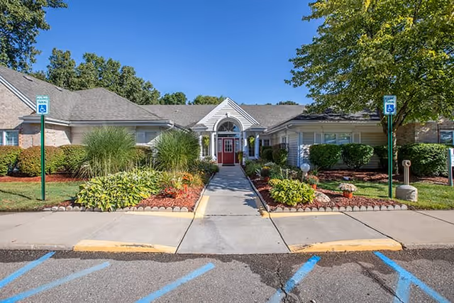 Front exterior view of a single-story building with a red door entrance, surrounded by landscaped bushes, plants, and trees under a clear blue sky. There are two handicap parking signs visible near the entrance.