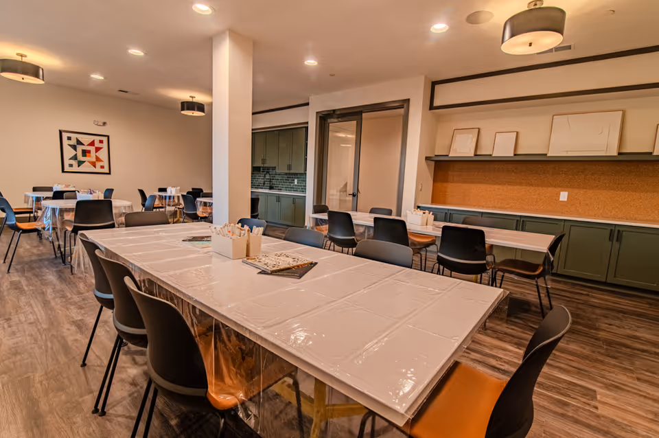 A spacious room with multiple tables covered in clear plastic tablecloths, surrounded by black and brown chairs. The room has wooden flooring, modern ceiling lights, and a wall with green cabinets and a corkboard above them. There are framed artworks resting on a shelf above the corkboard. In the background, there is a doorway leading to another room with green cabinetry and a tiled backsplash.